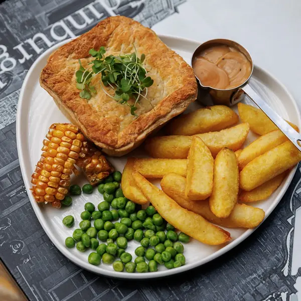 A burger on a dark plate with chips, corn ribs and garden peas