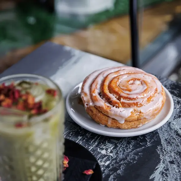 A hand holding a cinnamon swirl beside an iced coffee on a dark wooden table